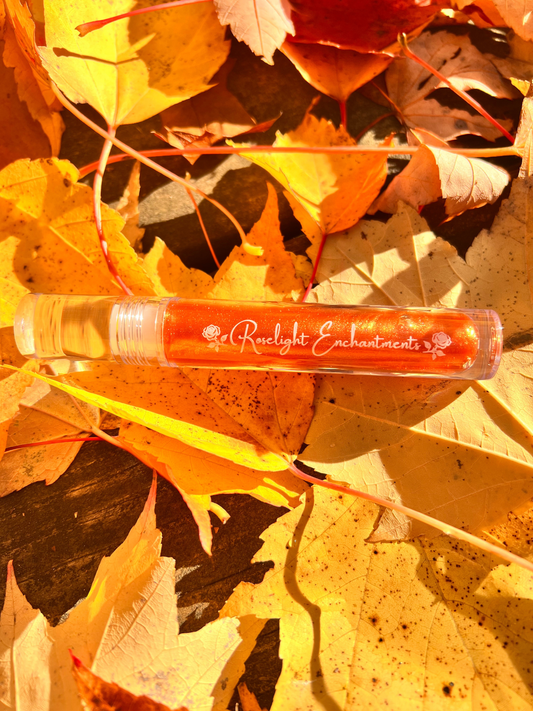 Lip gloss tube with "Roselight Enchantments" branding on a background of autumn leaves.